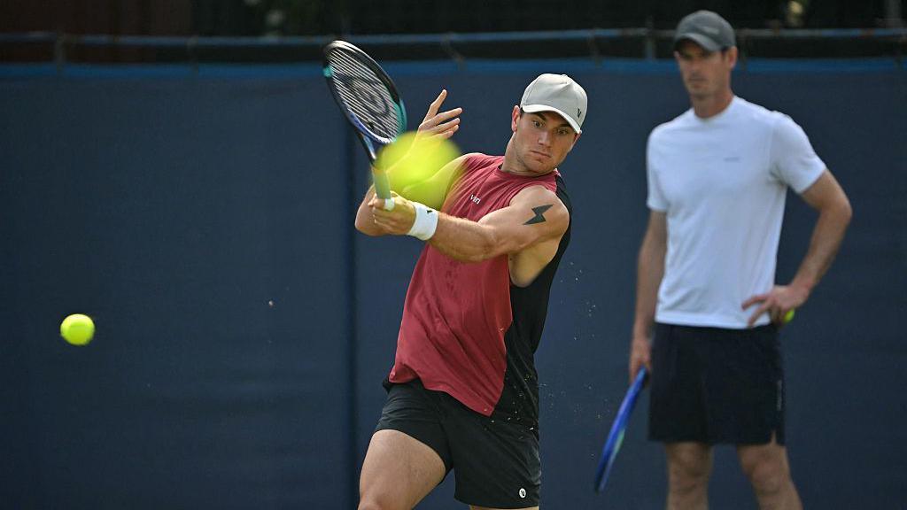 Jack Draper hits a forehand with Andy Murray in the background
