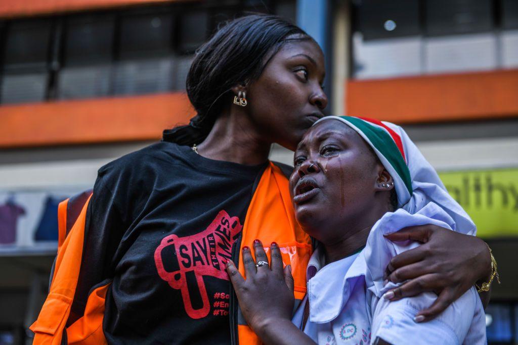 A woman embraces and comforts another woman who is crying. Both are at the protest in Nairobi. Behind them are shops.