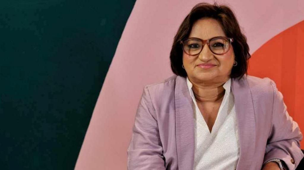 Samina Hussain smiling at the camera is wearing purple framed glasses and a purple jacket over a white blouse.