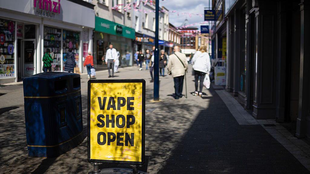 A high street scene in which there is a yellow sign with "Vape Shop Open" in black text. The sign is next to a bin. Pedestrians are in the background and there are shops either side of the walkway.