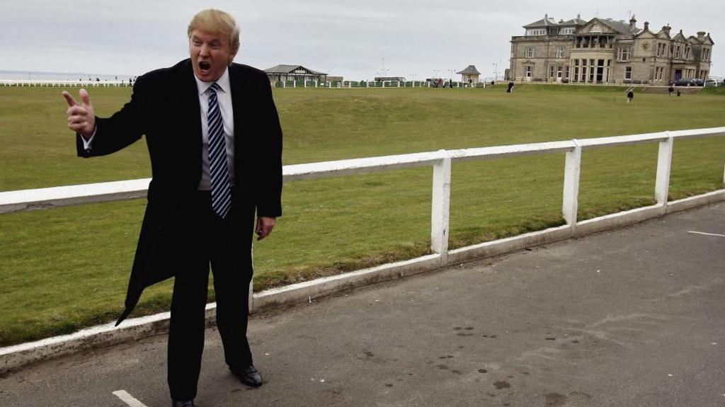 Donald Trump in black coat and suit with white shirt and blue and white striped tie, standing on the road by the side of the 18th fairway to the Old Course, St Andrews. He is pointing and shouting, in a pose associated with his TV programme The Apprentice. The Royal and Ancient building is behind him and the sky is grey.