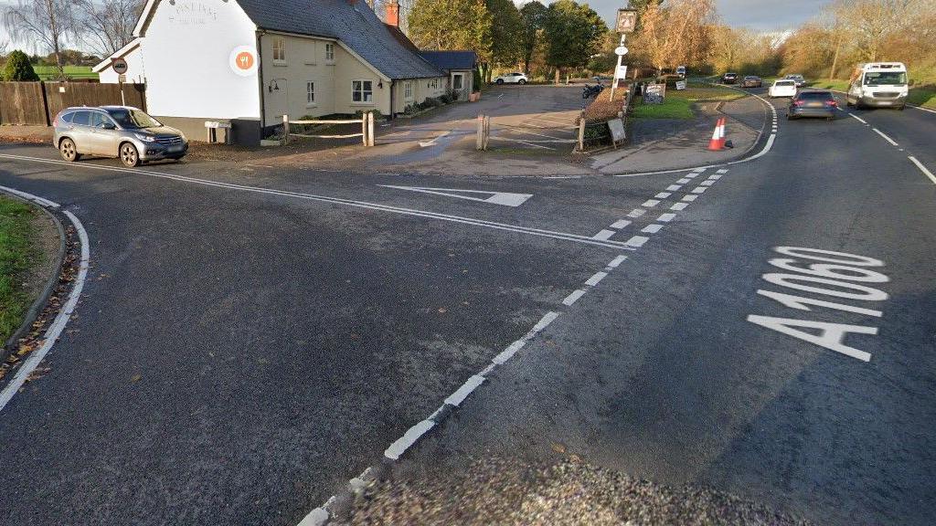 The junction of a main road with several vehicles travelling on it and a side road with one car on it. There is a large cream-coloured house on the corner and trees in the distance.