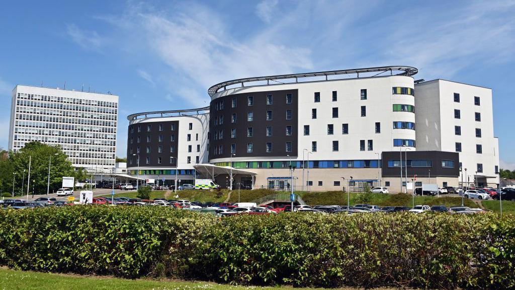 Victoria Hospital is a modern white and black building. It is surrounded by a car park and bushes.