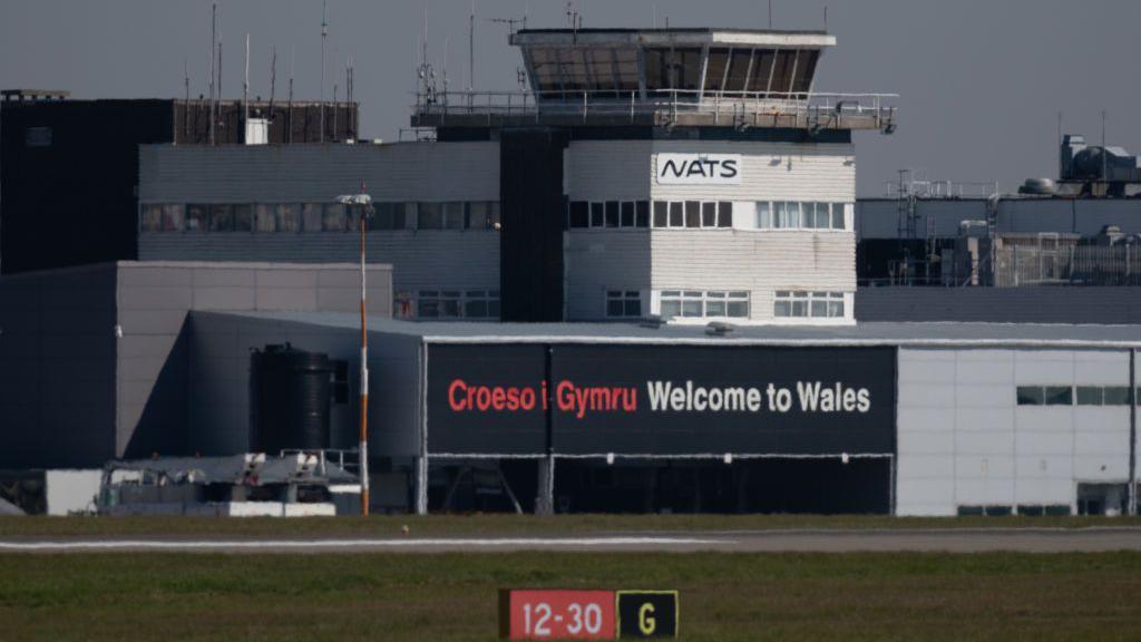 Adeiladau maes awyr Caerdydd, gyda'r geiriau "Croeso i Gymru" yn goch a "Welcome to Wales" yn wyn ar wal ddu.