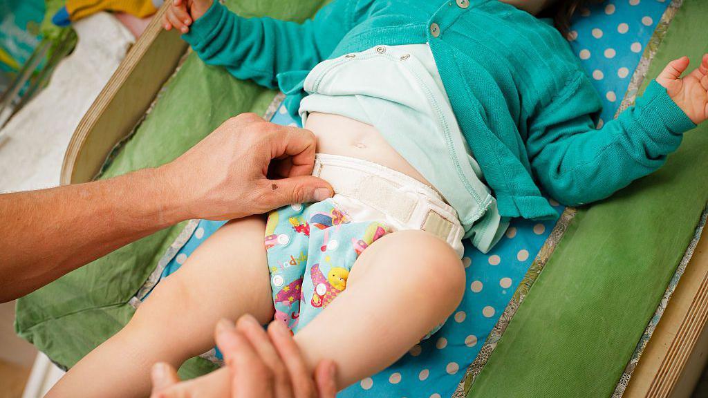 A baby on a green changing mat having their nappy changed. It's a reusable colourful cloth nappy