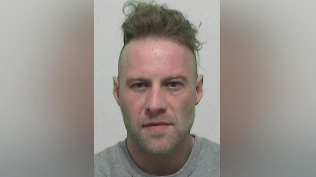 Mugshot of Henry. He has long brown hair stacked on top of his head with shaved sides and slight stubble around his chin.
