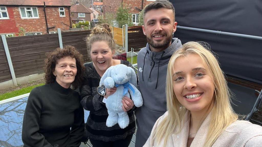 An older woman, two young women and a young man stand smiling at the camera as the young woman on the right takes a "selfie". The other young woman is holding a blue stuffed rabbit.