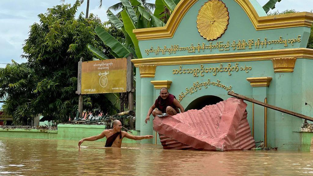 A Buddhist monk wades through flood waters as another sits on a broken roof in front of a monastery in Sin Thay village in Pyinmana, in Myanmar's Naypyidaw region, on September 13, 2024, following heavy rains in the aftermath of Typhoon Yagi.