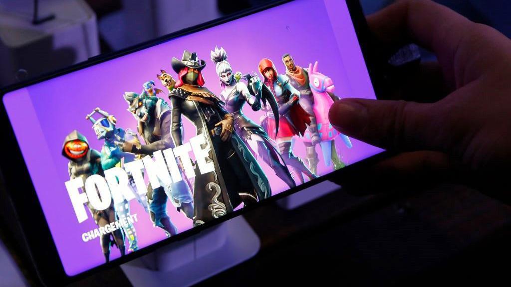 A person holds a Samsung smartphone that displays the loading screen of  Fortnite Battle Royale, which shows a mix of colourful playable characters from the game against a bright purple background.