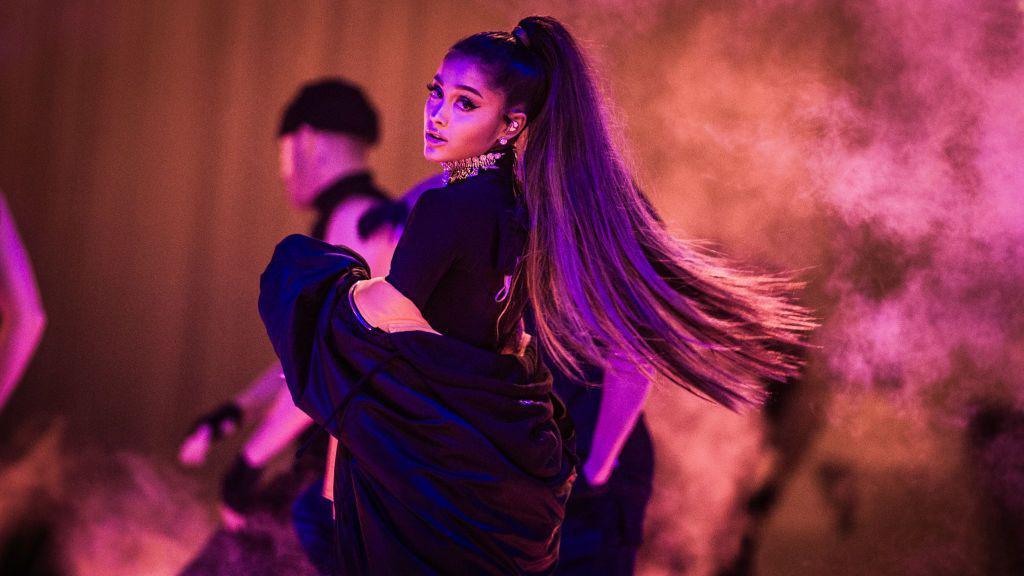 Ariana Grande performing on stage. She has long hair in a ponytail. She is wearing a oversized black coat over a black t-shirt.