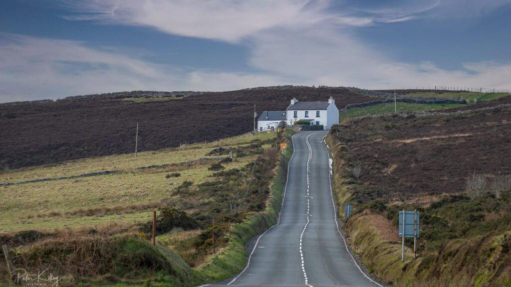 A long winding road with an embankment on the right, and green hills on the left, a white cottage is on the right of the road at the top. 