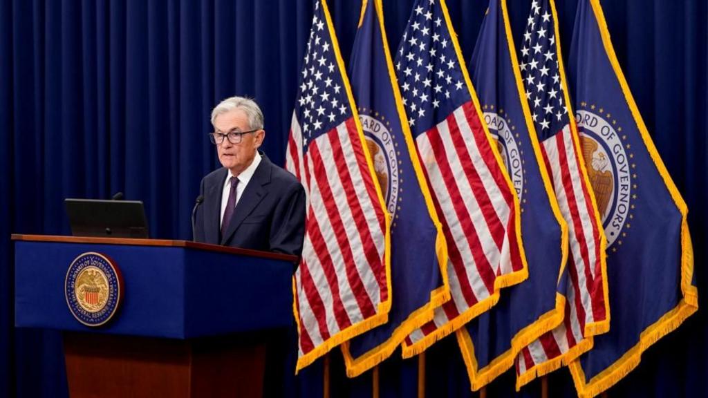 Jerome Powell, a man with grey hair and black square glasses, speaks at a podium in front of American flags and Federal Reserve flags, wearing a dark suit and purple tie