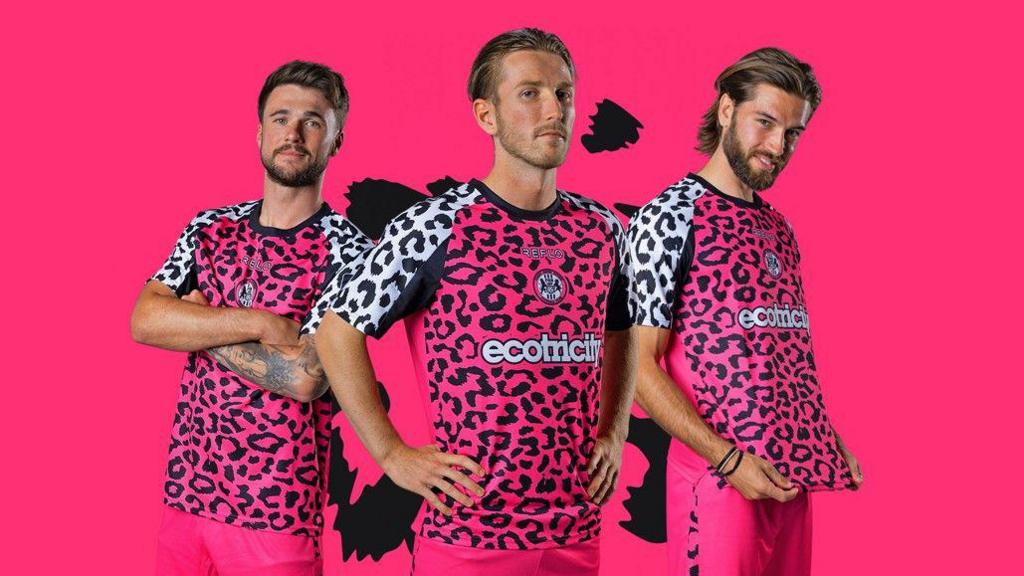 Three teammates of the Forest Green Rovers football club. They are posing for a promotional image against a bright pink background to showcase the new kit design. They are all wearing florescent pink shorts and matching tops, which are covered in a leopard print, with white sleeves. The man on the left has his arms folded, the one in the middle has his hands on his hips, and the one on the right is holding out the bottom hem and smiling.