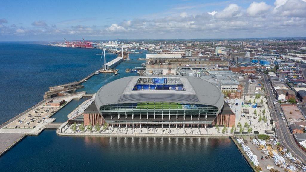 A large stadium sits on a dock with the River Mersey to the left and the outer city of Liverpool to the right