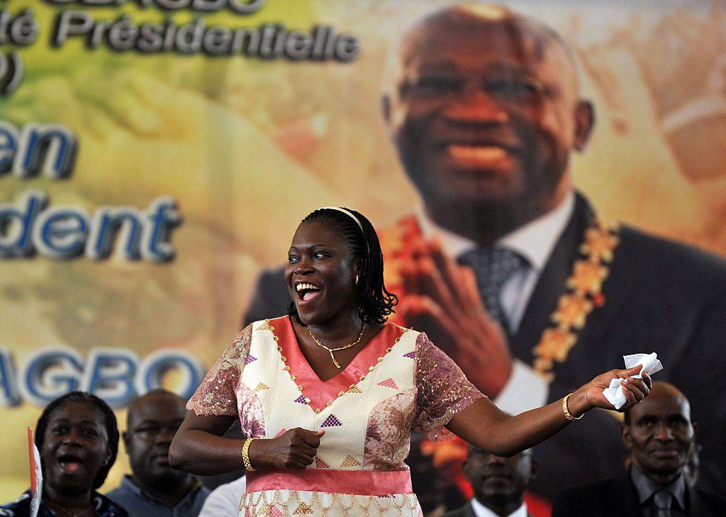Simone Gbagbo stands in front of a picture of her husband, wearing a pink and wife dress.