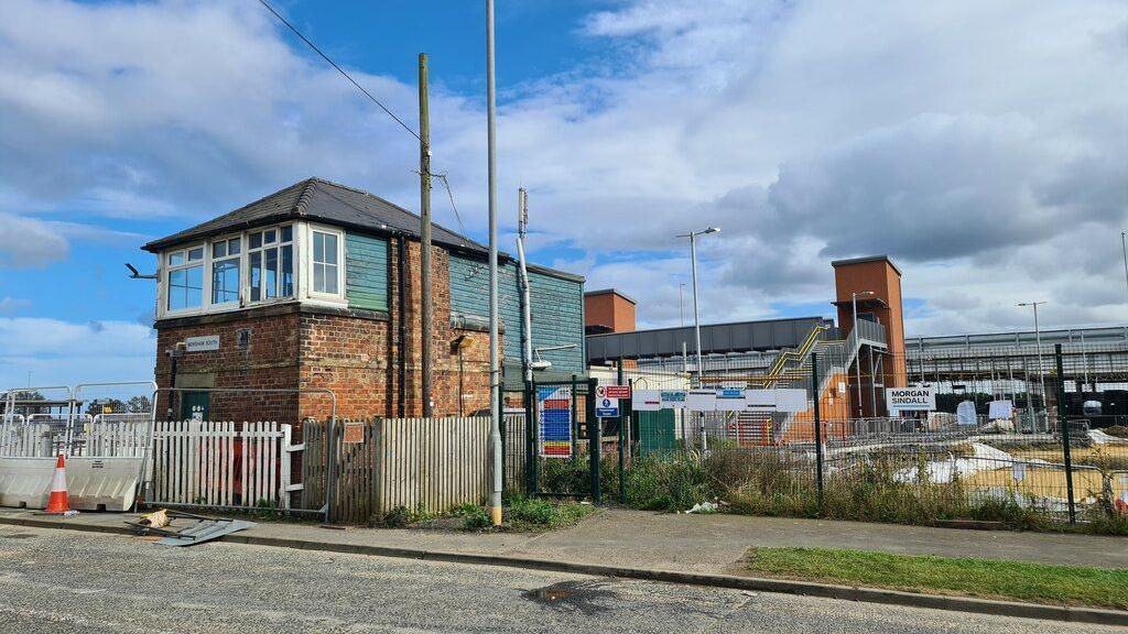 In the foreground Newsham signal box stands next to a road. It is a rectangular brick building with white framed windows overlooking the line. Behind there is the new lifts and passenger footbridge at the new station 