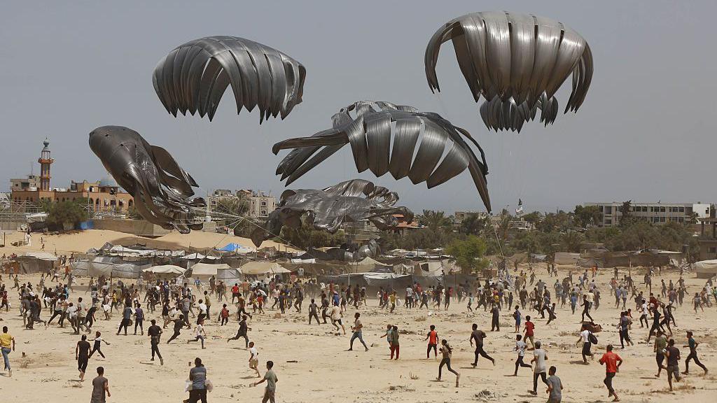 Black parachutes with crates of aid attached fall out of the grey sky onto a barren sandy landscape, with hundreds of people running to try and open them. 