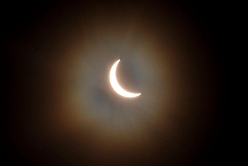 The sun is pictured during a partial solar eclipse. The sky is dark and cloudy. The sun is a thin arc in the sky.