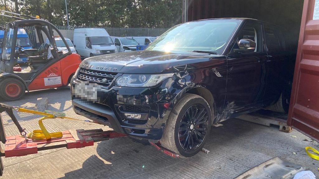 Image of the front of a black Range Rover with scratches on the side being pulled out of a red container. There are other vehicles in the background.