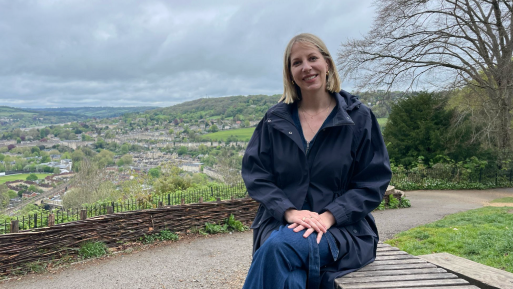 A woman sits on a park bench with her hand resting on top of her crossed legs. She is sitting alongside a park which overlooks Bath, with rooftops, fields and trees visible in the background. It is an overcast day with grey clouds filling the sky.