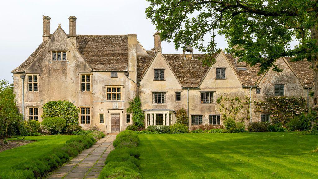 The front of a 16th century manor house - lots of pitched roofs, stone window frames and chimneys. A large neat lawn and pathway out front, with tree branches hanging over from the right
