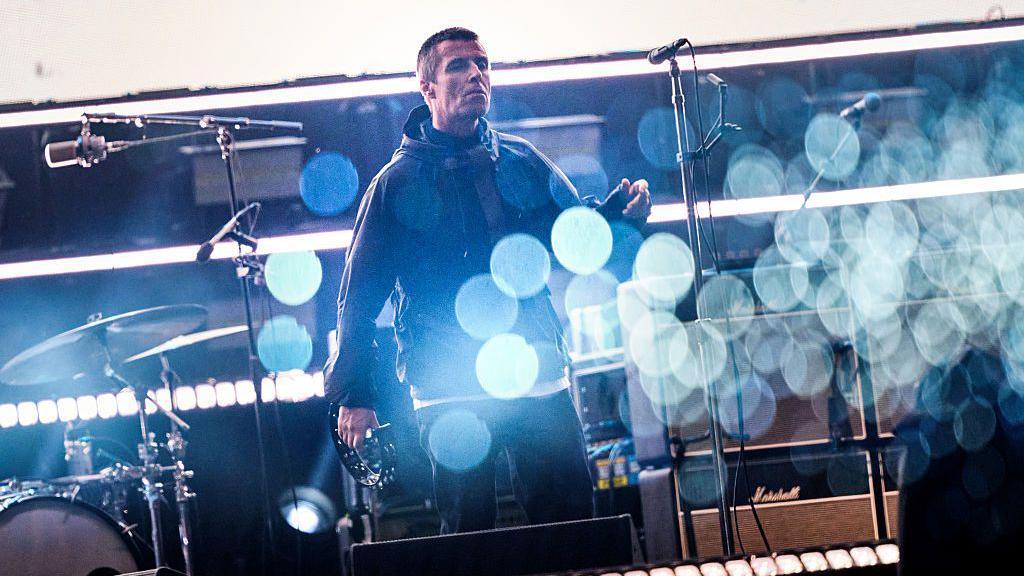Liam Gallagher stands on stage at Principality stadium. Glimmers of light create circles across the image. Liam wears a dark green parka. 