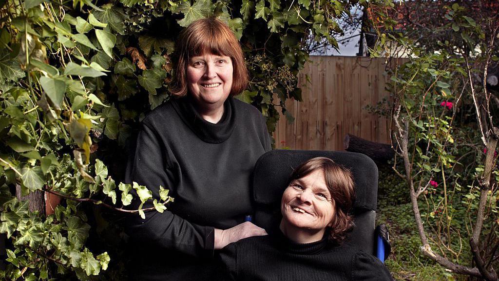 Rosemary Crossley, wearing a dark top and looking straight at the camera, and Anne McDonald in a wheelchair. The picture appears to be professionally taken and they are in a back garden.