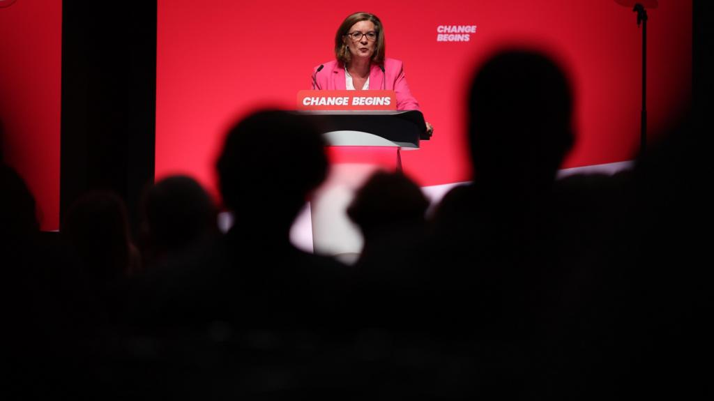 Eluned Morgan speaking at a Labour conference in front of a crowd of people who are in darkness.