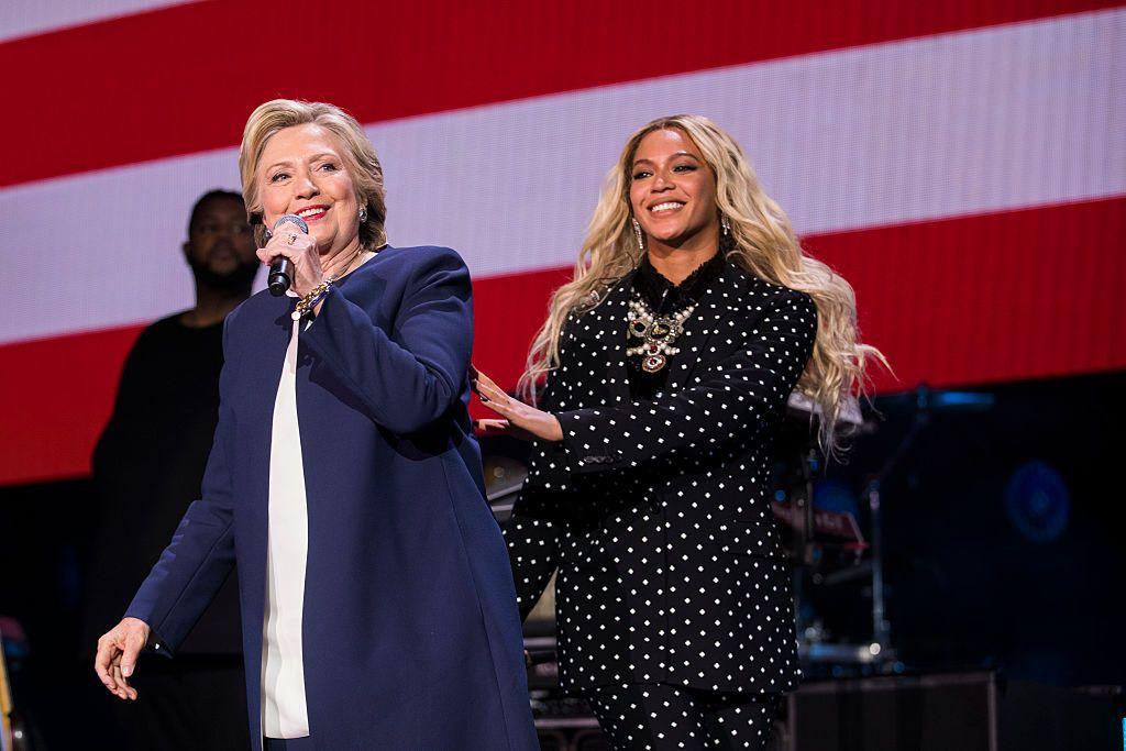 Hillary Clinton and Beyoncé appear on stage together at a rally in 2016. Clinton is speaking into a microphone while the singer is raising her hand towards Clinton's arm. Clinton wears a long, blue jacket. é has a black and white spotty suit on.