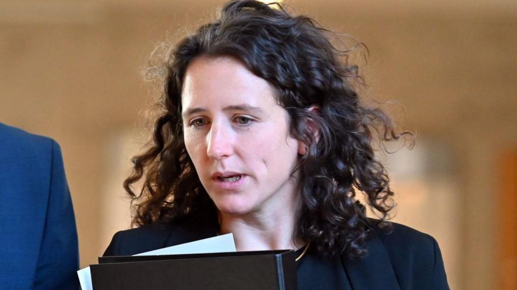 A woman with dark curly hair carrying a folder and papers 