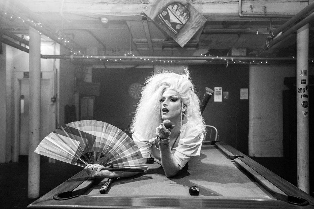 Image of a clubber with long, blonde hair holding a fan and a billiard ball, lying on a billiards table