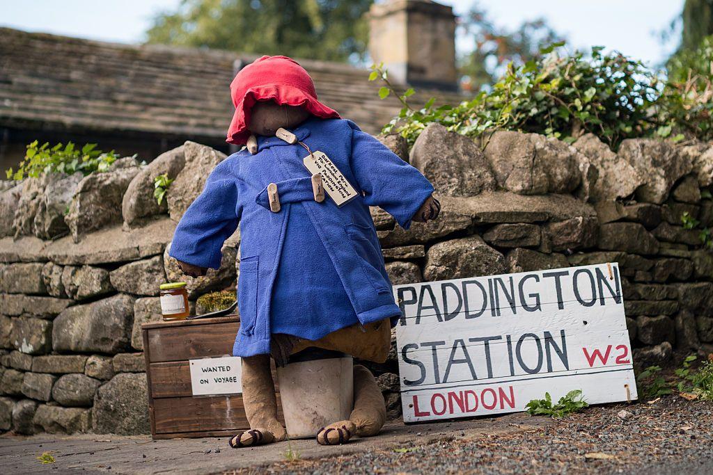 The scarecrow is standing in front of a brick wall, wearing a blue Paddington Bear jacket and red hat. On his right is a sign that says Paddington Station and to the left is a wooden box with a jar of marmalade on top.
