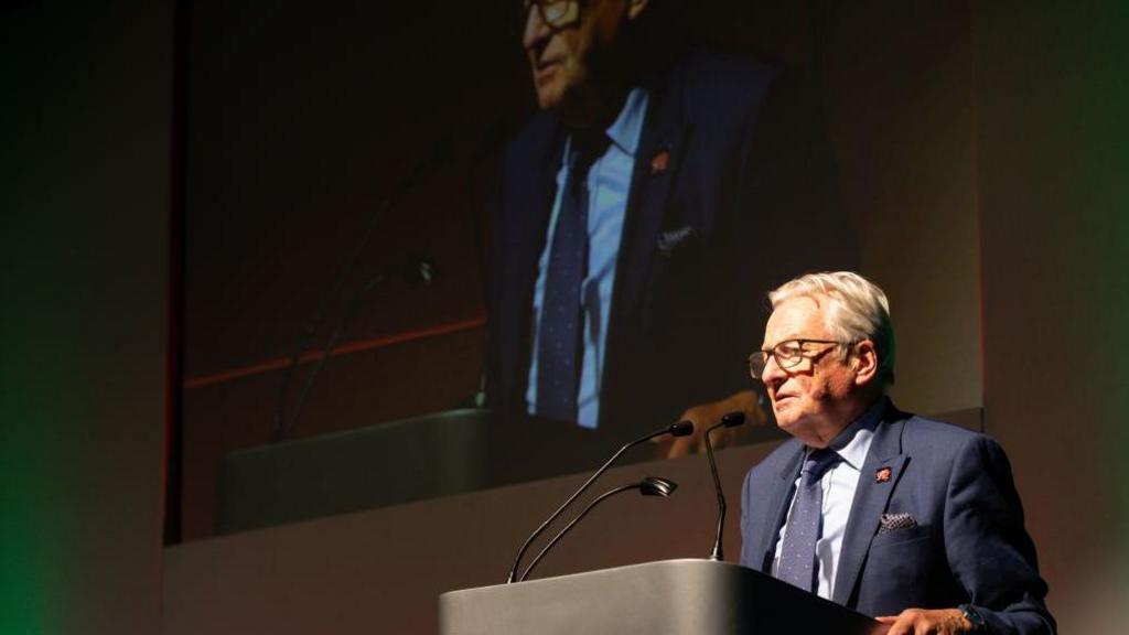 Dafydd Elis-Thomas giving a speech at a lecturn. His image can be seen projected behind him