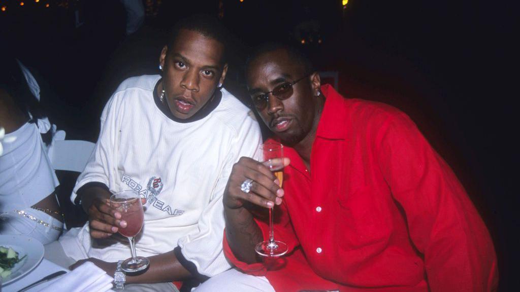 Jay-Z and Sean "Diddy" Combs at an event at Mr Combs' house in 2000. Jay-Z sits on the left at a table holding a beverage. Mr Combs sits next to him, also holding a drink.