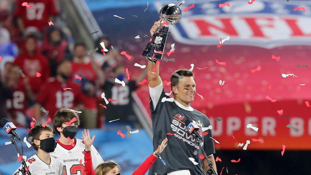 Tom Brady holds aloft the Vince Lombardi trophy with red and white confetti falling, next to three children, all wearing Covid-19 facemasks 