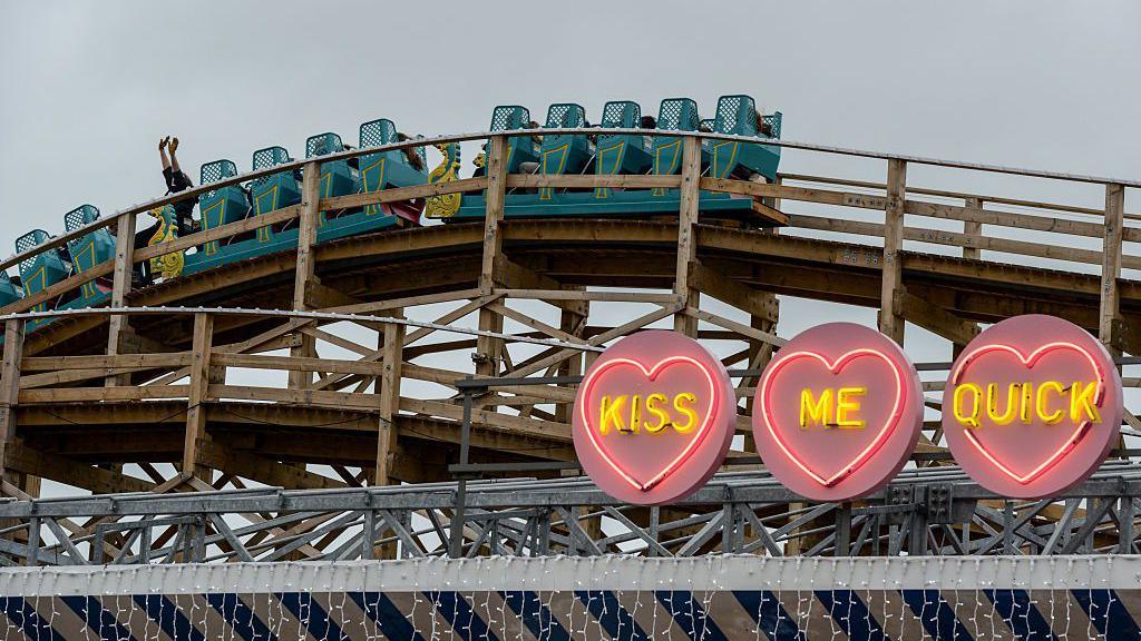 Një person në një karrocë me rollercoaster blu me krahët lart ndërsa makinat kalojnë mbi një hark prej druri. Përpara rollercoaster-it ka tre drita zemre neoni. Në secilën zemër janë fjalët "kiss", "me" dhe "quick".