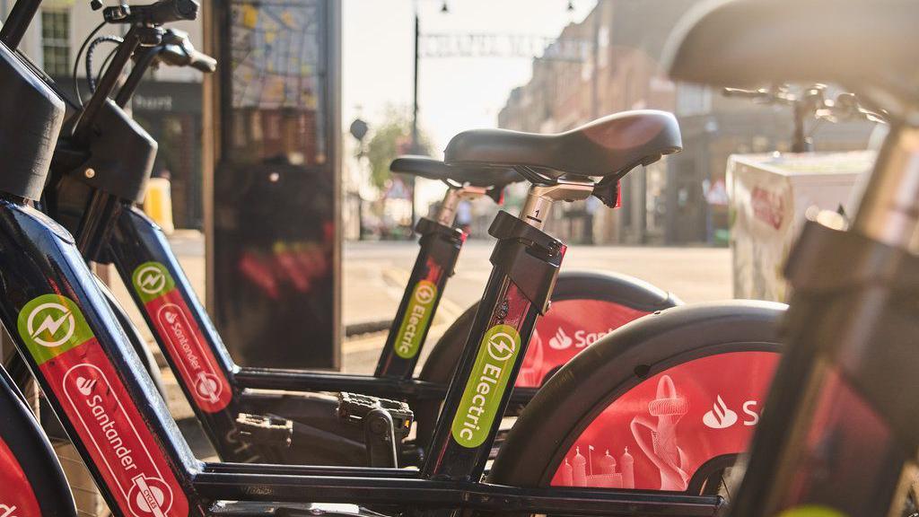 A row of red Santander electric bikes is neatly parked at a docking station on a city street. Behind them, there's a map display and buildings lining the pavement.
