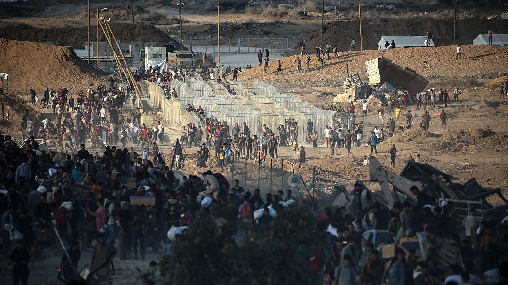 Hundreds of people queue near metal fences on a barren, arid landscape for food. 