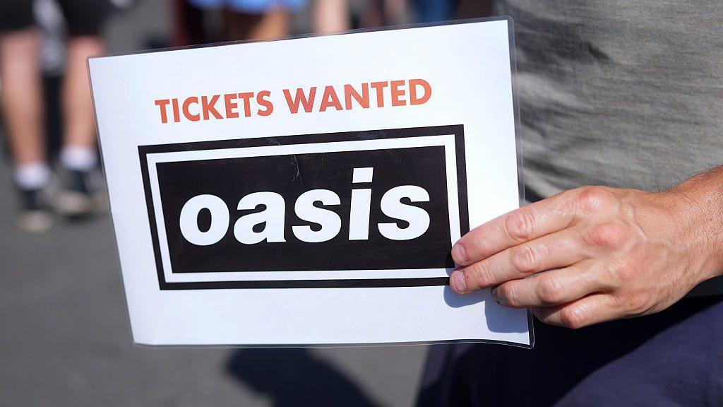 A mans hand holds a piece of laminated white paper which reads 'Tickets wanted' in red font and a black box with white righting that reads 'Oasis.' The man holding the sign has on navy trousers and a grey top.