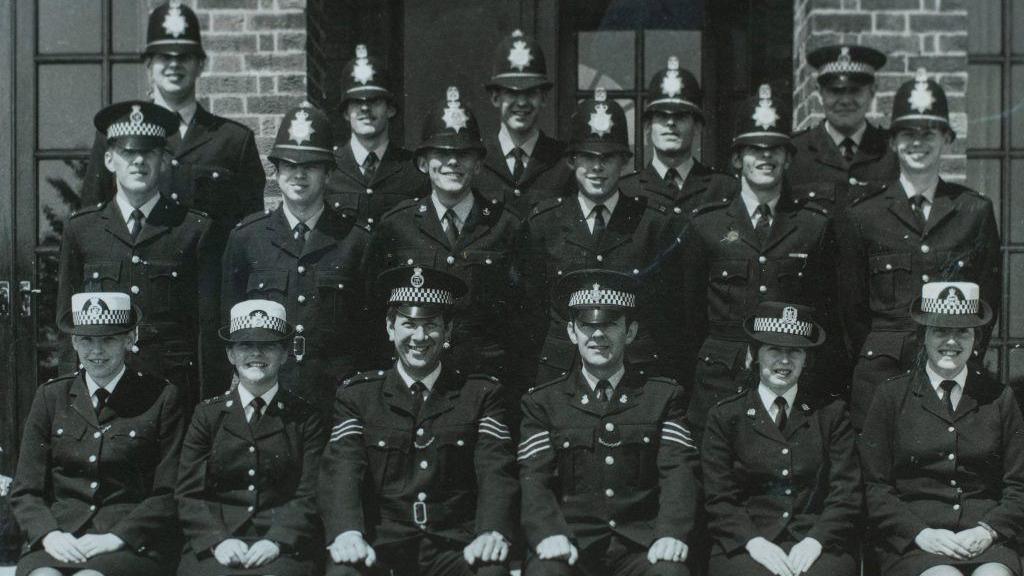 A three rows of police officers, the image is in black and white.
All the officers are wearing uniform, with four women sat in the front row. They are all looking at the camera.