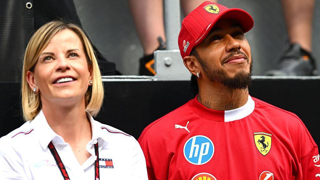 Susie Woolf in white shirt is standing smiling and look up at something. To her right, Lewis Hamilton wears a red cap and shirt with Ferrari branding. He too is looking up at something and smiling.