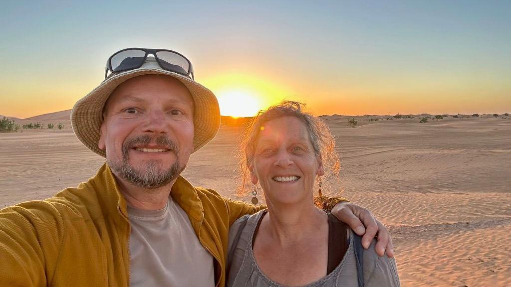 A selfie taken by Nick Smith showing him wearing a yellow jacket and a beige undershirt, and a beige hat with sunglasses sitting on top of his head, his arm around his wife, who is smiling and has an updo and is wearing dangling earrings. Behind them is the sun setting over the Moroccan desert. 