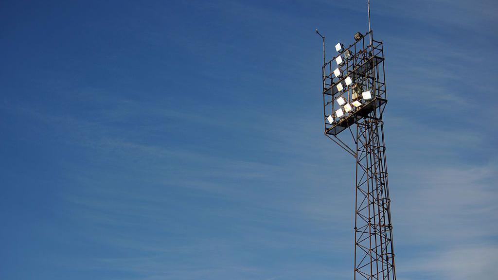 A large floodlight at Rockingham Road stadium. It is partly lit against an early evening sky.