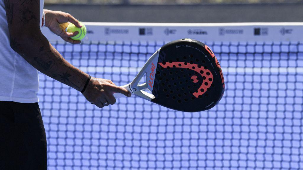 Image shows a black padel tennis racket and tennis ball being held in front of a net on a tennis court 