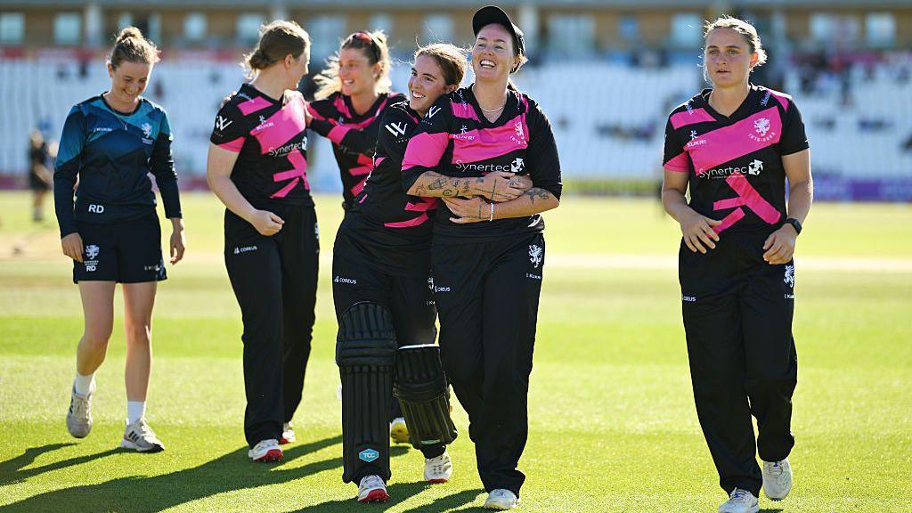 Women in cricket kits are walking on a green field hugging each other and laughing.