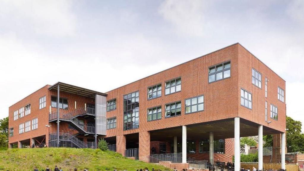 Modern buildings at Moseley School with children in uniform outside in the playground. The building is on columns with an open area underneath and grassy areas around it.