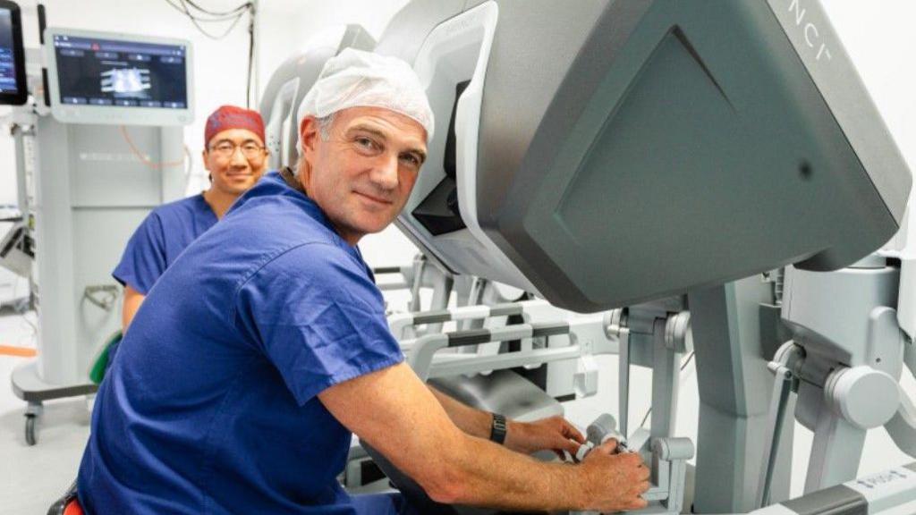 Kyle is in the foreground wearing a health uniform and sitting next to equipment. Mr Mak is in the background.