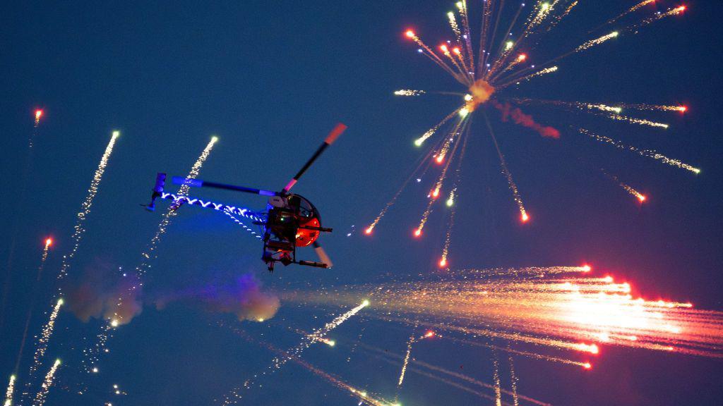 A helicopter with blue lights on its tail flies in a dark sky, with red fireworks going off around it.