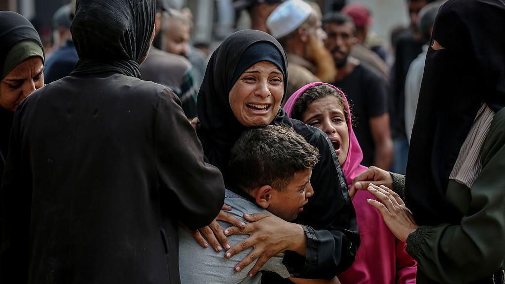 A crying woman wearing black hugs a young boy, with other crying people around them. 