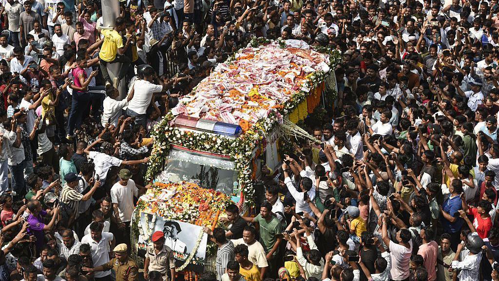 People gather as the mortal remains of singer Zubeen Garg are taken to his residence from the Guwahati airport on 21 September 2025, in a garlanded vehicle with a poster of the musician on the bonnet.

